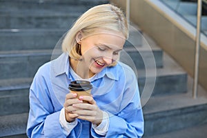 Close up portrait of beautiful, smiling blonde woman, student sitting on stairs outside campus, drinking takeaway coffee
