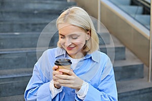 Close up portrait of beautiful, smiling blonde woman, student sitting on stairs outside campus, drinking takeaway coffee