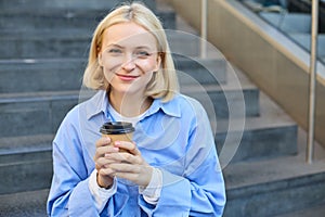 Close up portrait of beautiful, smiling blonde woman, student sitting on stairs outside campus, drinking takeaway coffee