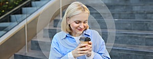 Close up portrait of beautiful, smiling blonde woman, student sitting on stairs outside campus, drinking takeaway coffee