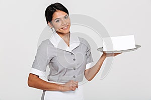 Close-up portrait of beautiful smiling woman in gray uniform holding metal tray with empty sign card, looking at camera