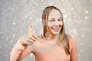 Close up portrait of beautiful smiling young woman in a beige shirt looking and showing thumb up, on a background of gray sparkles