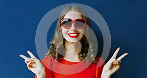 Close up portrait of beautiful smiling young woman wearing a red heart shaped sunglasses over a blue
