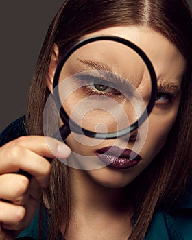 Close-up portrait of beautiful woman with dark lips posing, holding magnifying glass over dark grey background. Perfect