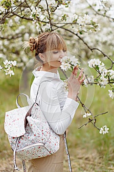 Close-up portrait of beautiful young blonde woman in white shirt with backpack posing under apple tree in blossom in Spring