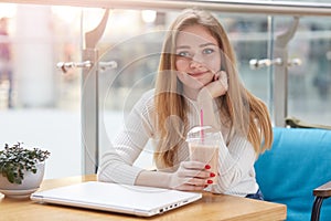 Close up portrait of beautiful young caucasian woman with long blonde hair sitting in cafe, drinking milk cocktail, having rest