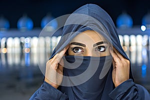 Close up portrait of beautiful young Muslim woman wearing Hijab looking away with Mosque at background during night