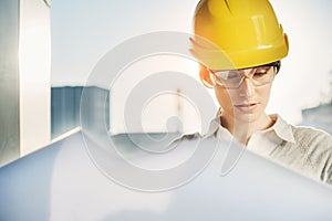 A beautiful young woman engineer is in front of a construction metal wall wearing protective gear, helmet and glasses.