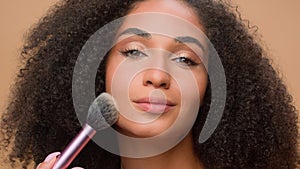 Close up portrait beige studio background beautiful African American woman with perfect smooth skin apply powder looking