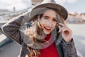 Close-up portrait of blissful blue-eyed girl touching glasses and laughing. Fashionable woman in gray hat and trendy
