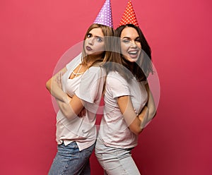 Close up portrait of blonde and brunette young women with birthday hats having fun isolated on pink background
