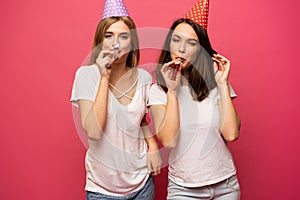 Close up portrait of blonde and brunette young women with birthday hats having fun isolated on pink background