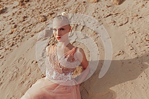 Close-up portrait of a blonde woman posing in embroidery dress in the desert, lying on golden sand with closed eyes.