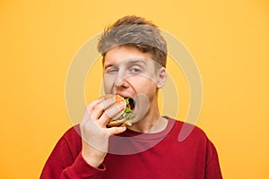 Close up portrait of a boy eats a burger on a yellow background and looks at the camera, young man bites a burger. Fast food