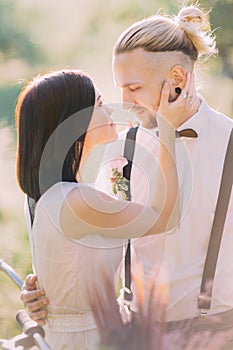 The close-up portrait of the bride in the white wedding dress petting the modern-dressed groom in the sunny spring