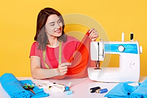 Close up portrait of brunette European female seamstress with tapes mesure on neck, wears casual red t shirt, isolated over yellow