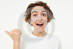 Close up portrait of brunette girl gasps and looks surprised, points left to inquire about promotion, stands over white