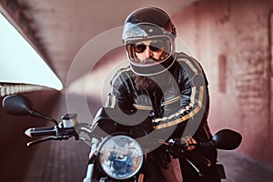 Close-up portrait of a brutal bearded biker in helmet and sunglasses dressed in a black leather jacket sitting on a