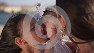 Close-up portrait of carefree pretty baby girl with cheerful sisters kissing toddler on cheeks in slow motion. Happy