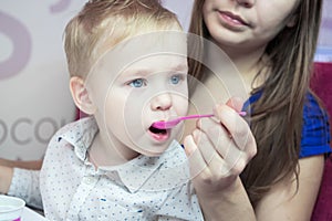 Close-up portrait of a caucasian kid boy with a big blue eyes, which his mother with a spoon is feeding ice cream in a cafe