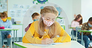 Close up portrait of Caucasian pretty joyful teen girl pupil in face mask studying at school writing while sitting at