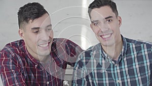 Close-up portrait of Caucasian twin brothers looking at camera and smiling. Two positive young men posing at home