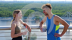 Close up portrait of Caucasian young sportswoman speaking with handsome sportsman outside in city with bottle of water