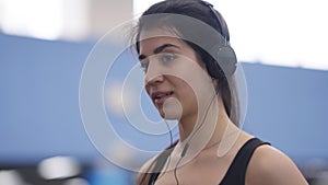 Close-up portrait of charming beautiful Middle Eastern sportswoman enjoying music in headphones walking on treadmill in