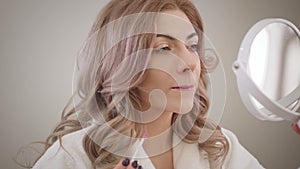 Close-up portrait of charming Caucasian girl looking at small round mirror and applying lipstick. Confident young woman