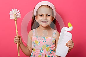 Close up portrait of charming girl holds detergent and plunger in hands, little cute housewife wearing hairband and sundress,