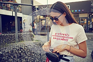 Portrait of a girl in dark sunglasses posing near a city mall. Dressed in white t-shirt, blue trousers, black waist bag