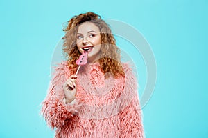 Close up portrait of cheerful smiling beautiful brunette curly girl in pink fur coateating lollipop over blue background