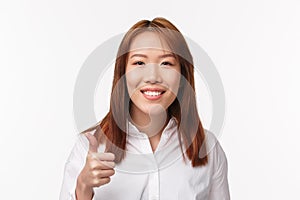 Close-up portrait cheerful smiling pleased asian woman in white shirt, say good job, nice work, make thumb-up gesture in