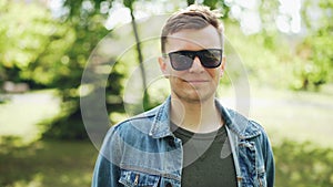 Close-up portrait of cheerful young man in sunglasses wearing denim jacket looking at camera and smiling. Sunny summer