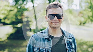Close-up portrait of cheerful young man in sunglasses wearing denim jacket looking at camera and smiling. Sunny summer