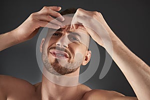 Close up portrait of concentrated frowning young bearded man plucking his eyebrows