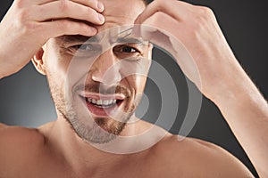 Close up portrait of concentrated frowning young bearded man plucking his eyebrows