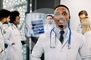 Close up portrait of confident African male doctor in front of his multiethnic medical team, looking at camera. Happy