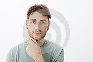 Close-up portrait of confident handsome mature male with fair hair in earrings, touching beard gently and smiling with