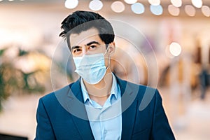 Close-up portrait of confident young man in stylish suit putting protection face mask looking at camera in hall of mall