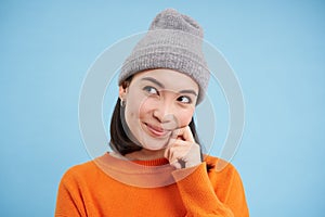 Close up portrait of coquettish asian girl in hat, looks aside and smiling, fantasizing, dreaming of smth, standing over