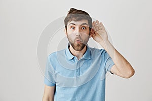 Close-up portrait of curious funny young man with beard and messy hair, holding hand near ear as if overhearing rumor