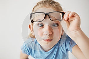 Close up portrait of curious little girl with big blue eyes standing close and looking in camera, holding glasses with