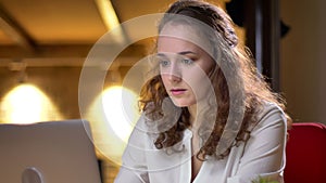 Close-up portrait of curly-haired businesswoman attentively working with laptop and smartphone simultaneously in office.