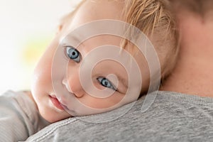 Close-up portrait of cute adorable blond caucasian toddler boy on fathers shoulder indoors. Sweet little child feeling safety on