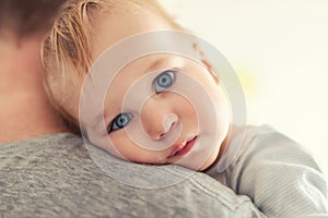 Close-up portrait of cute adorable blond caucasian toddler boy on fathers shoulder indoors. Sweet little child feeling safety on