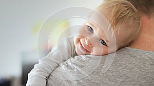 Close-up portrait of cute adorable blond caucasian toddler boy on fathers shoulder indoors. Sweet little child smiling