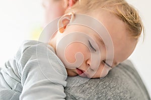 Close-up portrait of cute adorable blond caucasian toddler boy sleeping on fathers shoulder indoors. Sweet little child feeling