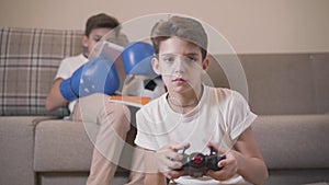 Close-up portrait of cute Caucasian boy playing game console as his twin brother sitting at the background in boxing