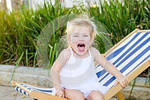Close up portrait of cute emotional blondy toddler girl in white dress sitting on the deckchair and yelling. City park recreation